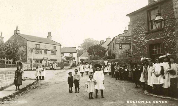 The image shows a group of children looking at the camera with old houses behind them. The photo dates from around 1900 and so the girls are dressed with smocks and hats and the boys in long shorts and caps. The old house behind them to the left dates from the late 1600s and is stone built with three floors and mullioned windows. It is a large house.